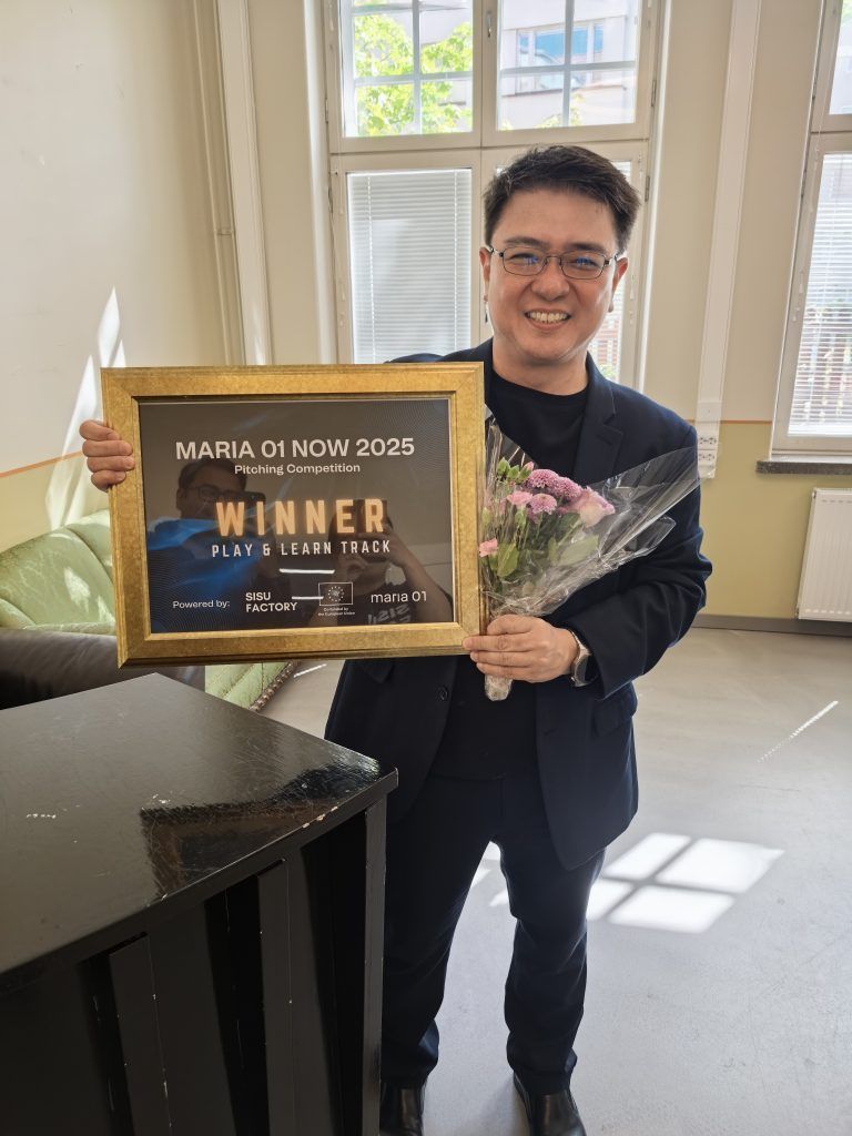 Same PSI representative smiling indoors, holding the framed "Maria 01 NOW 2025 Winner, Play & Learn Track" certificate and a bouquet of flowers