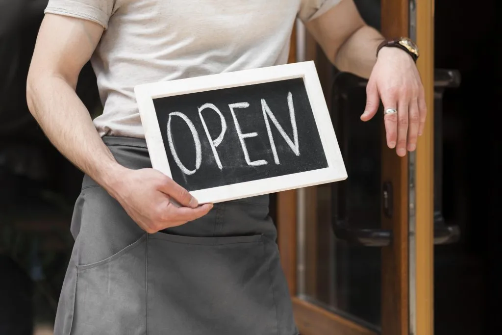 Stock photo of a person in an apron holding a chalkboard 'OPEN' sign while standing in a shop doorway