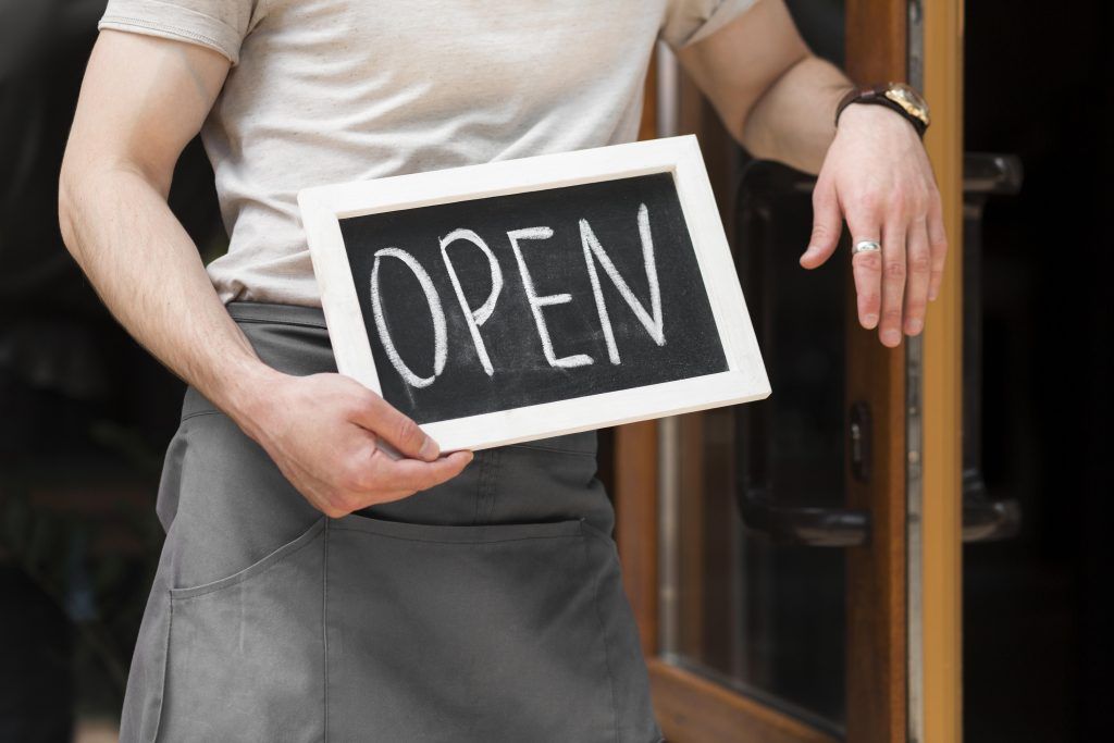 Stock photo of a person in an apron holding a chalkboard 'OPEN' sign while standing in a shop doorway