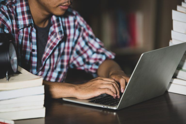 Stock photo of a student typing on a laptop at a table stacked with books in a library