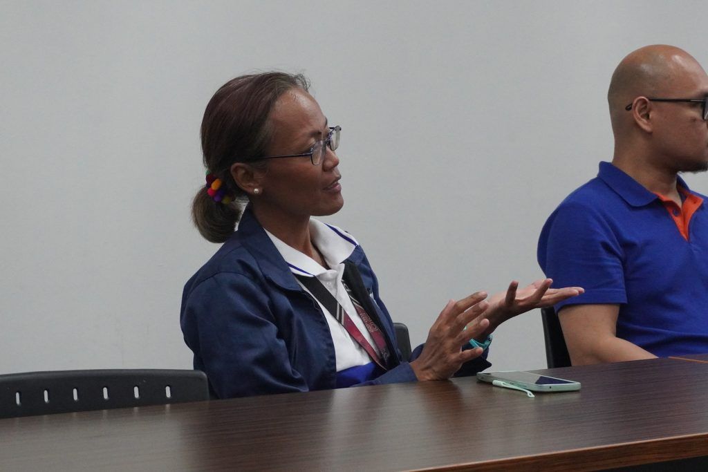 A PSHS teacher gesturing while speaking, seated beside a colleague in a meeting room