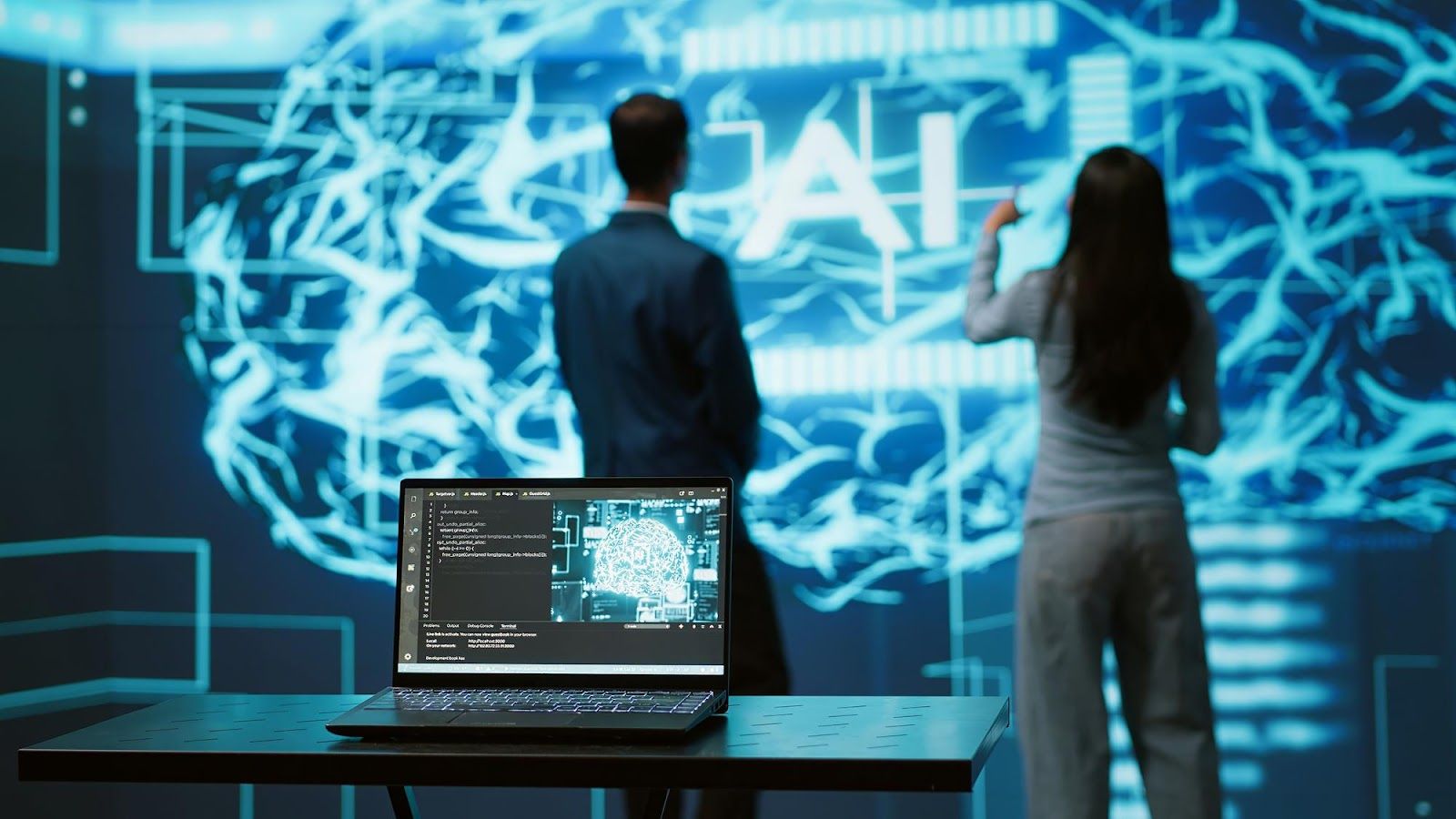 Stock photo of two people silhouetted before a glowing blue digital brain visualization labeled AI, with a laptop showing code and a brain graphic in the foreground