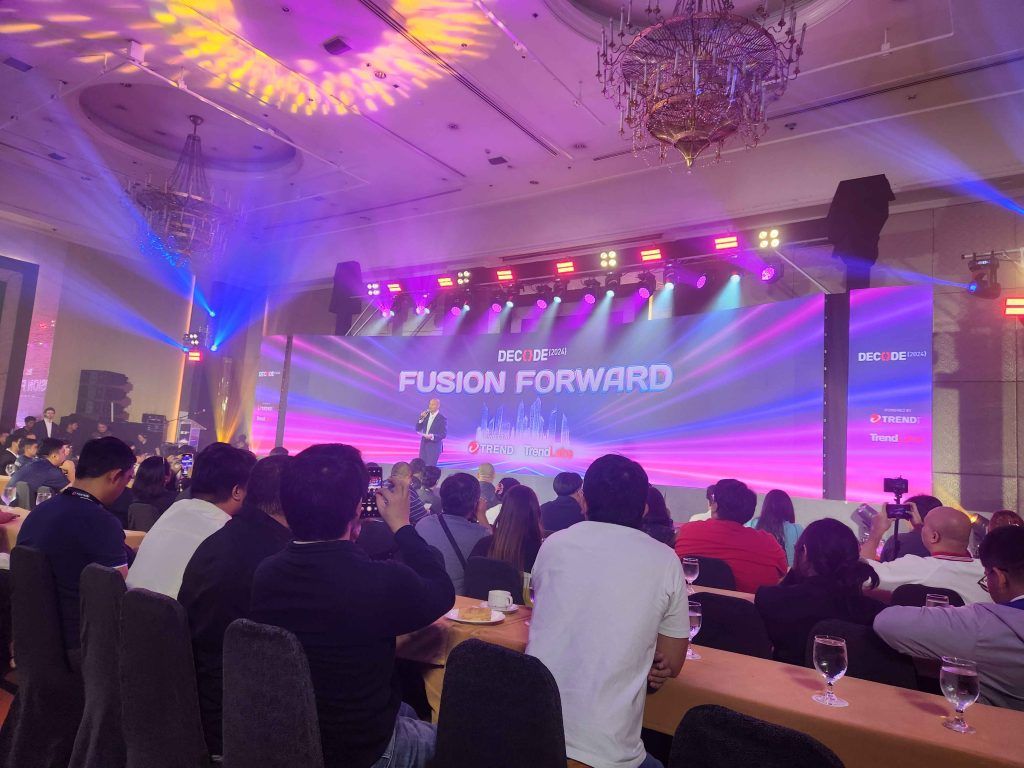 Audience seated at tables watching a speaker on stage under a large screen reading 'DECODE 2024: FUSION FORWARD' with colorful stage lighting