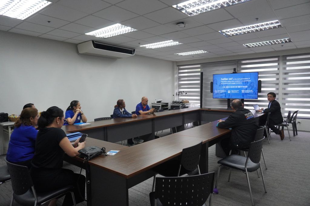 Wide shot of a meeting room where PSHS staff sit around tables watching a presentation on a wall-mounted screen showing the better-ed platform