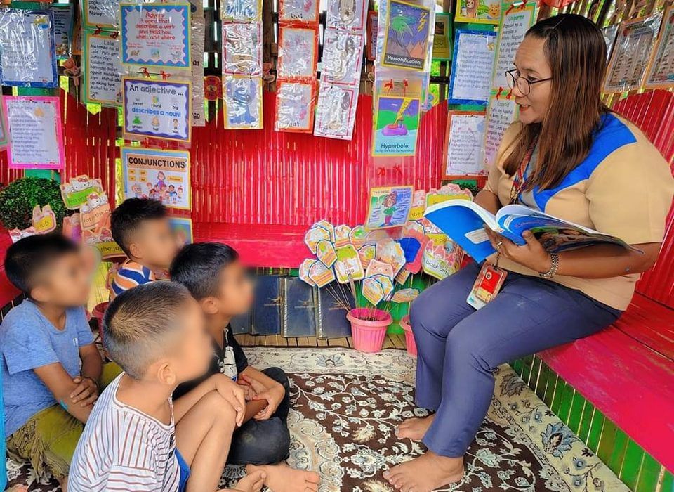Photo of a teacher reading a book aloud to young students seated in a brightly decorated classroom reading corner covered with vocabulary posters, credited to the Department of Education