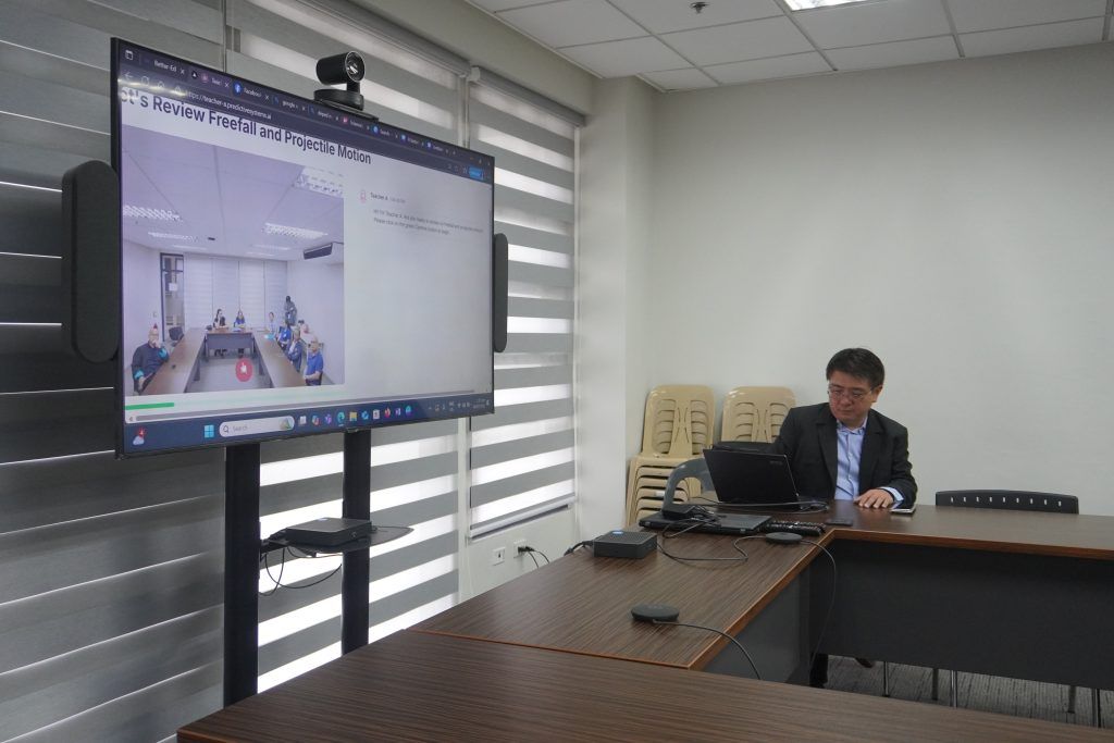 A presenter seated at a laptop beside a large screen displaying the better-ed platform mid-demo, showing a video call interface and a chat titled 'Review Freefall and Projectile Motion'