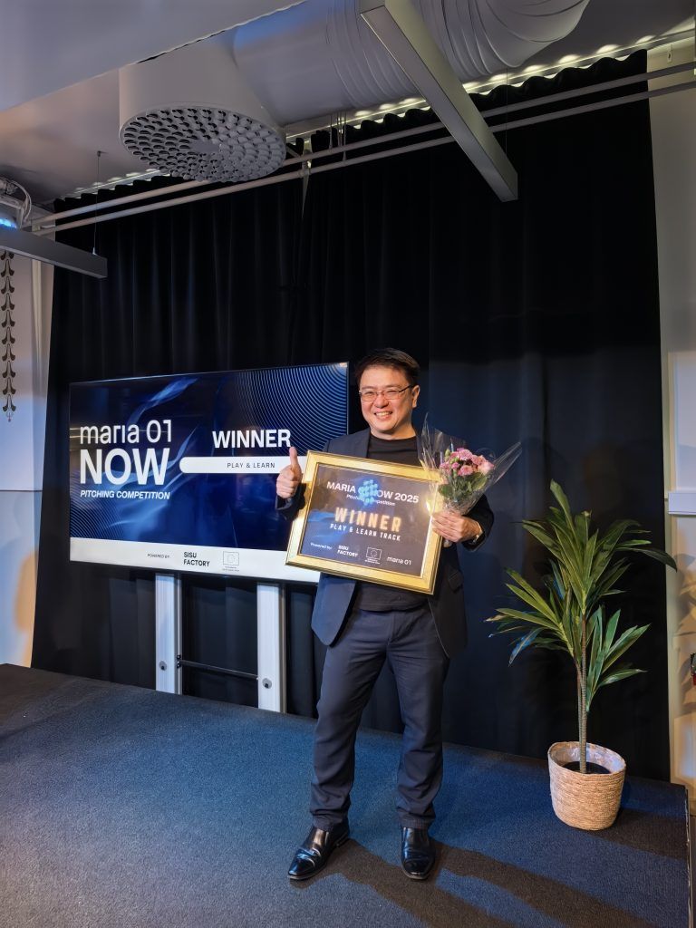 PSI representative on stage giving a thumbs up while holding a bouquet and a framed "Maria 01 NOW 2025 Pitching Competition - Winner, Play & Learn Track" certificate, with the award screen displayed behind him
