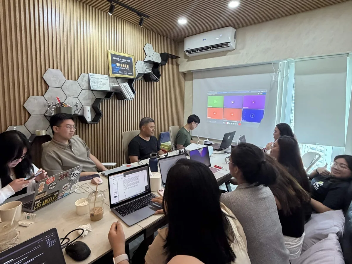 The team at a long desk during a standup, laptops open, a shared board projected on the wall behind them.